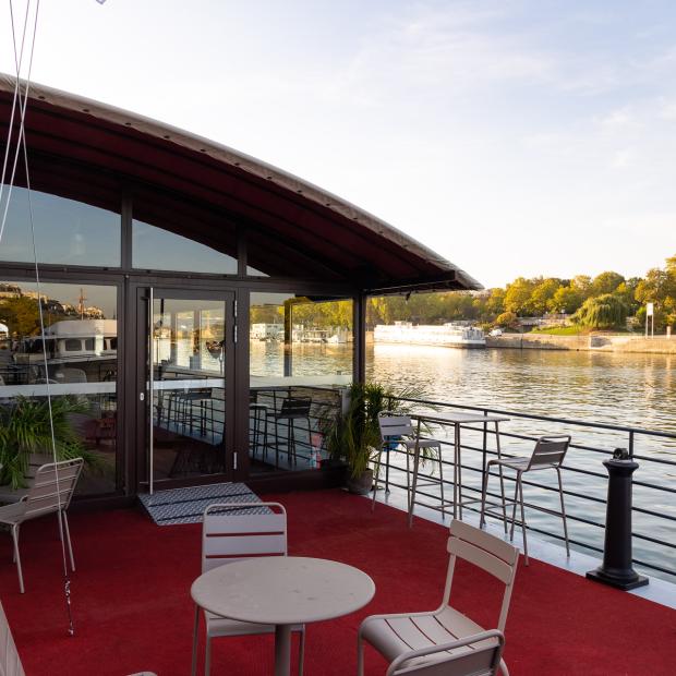 Terrasse extérieure d'un bateau restaurant sur la Seine, tables et chaises beiges, vue sur le fleuve