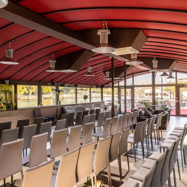 Salle de conférence sur péniche à Paris, rangées de chaises grises, plafond rouge, vue sur la Seine