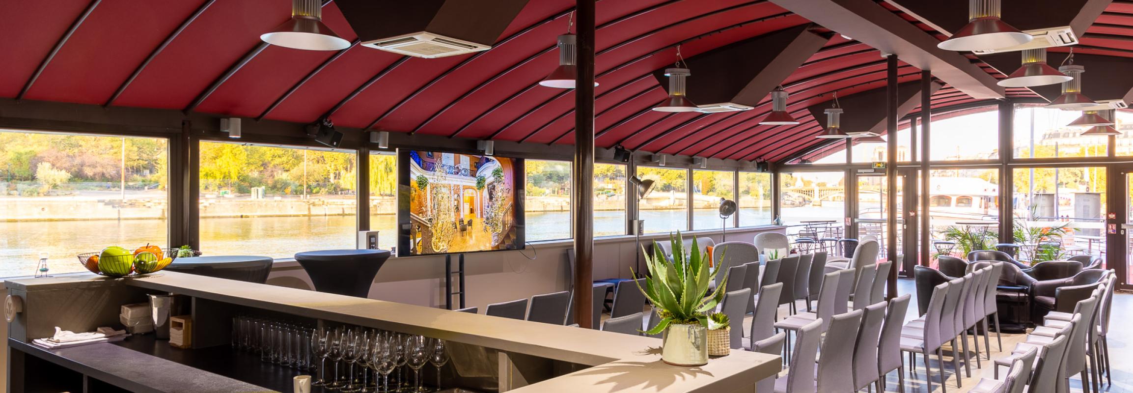 Salle de réception intérieure péniche Paris avec bar, chaises grises et vue sur la Seine